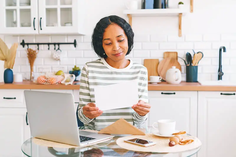 Woman reading mail with computer open on a glass kitchen table. She is smiling and the kitchen is all white with wood accents.