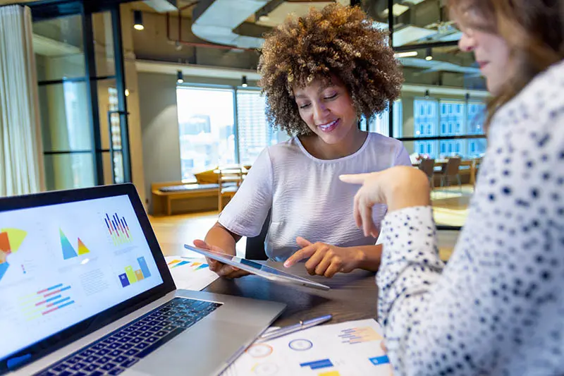 Women in a modern office reviewing graphics and charts, reviewing direct mail marketing tips on a laptop and tablet