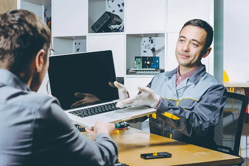 A computer repair shop employee wearing latex gloves is handing a MacBook to a customer thanks to Direct Mail. 