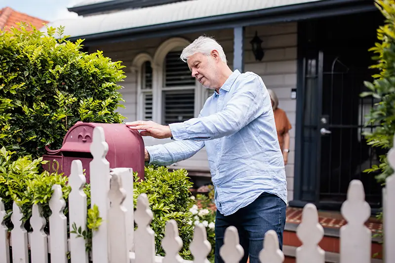 a man in his 60s opening a mailbox with a white picket fence 