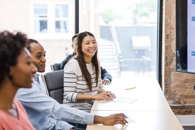 Modern Postcard’s Direct Mail Crash Course, Sponsored by the USPS® - A group of people at a table attending a seminar while taking notes and smiling